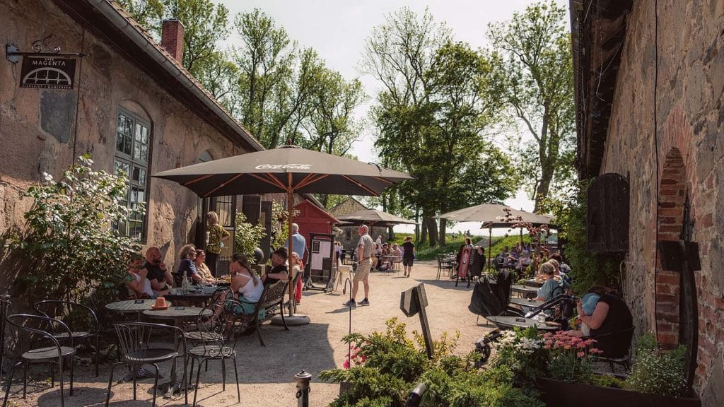 Folk nyter mat og drikke i solveggen ved Café Magenta på Bastion 5 i Gamlebyen i Fredrikstad. Uteservering under parasoller og blomstrende omgivelser.