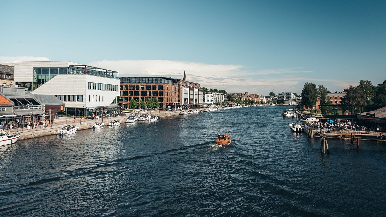 Bryggepromenaden i Fredrikstad sentrum med båter, moderne bygg og folk som nyter sommerdagen ved vannet.