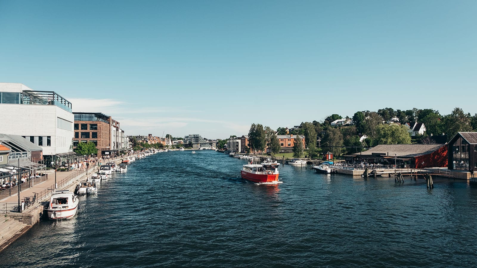 Byferga på elva i Fredrikstad sentrum, omgitt av småbåter, brygge, og folk på uteservering.