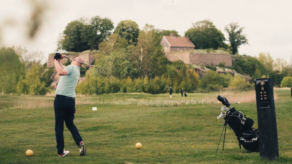 Golfspiller slår ut på banen ved Kongsten fort, Gamle Fredrikstad Golfklubb, med golfbag og historiske murer i bakgrunnen.