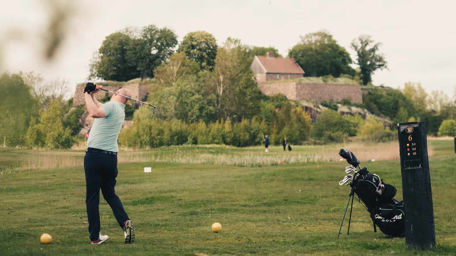 Golfspiller slår ut på banen ved Kongsten fort, Gamle Fredrikstad Golfklubb, med golfbag og historiske murer i bakgrunnen.