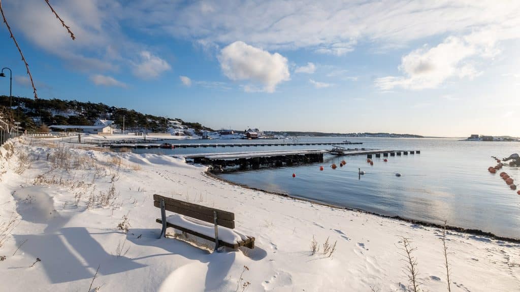 Snødekt strand med benk og brygger ved Kroksand på Hvaler en klar vinterdag. Svaner svømmer i det stille vannet under en blå himmel med lette skyer.