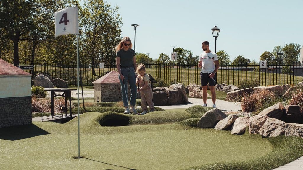 Barn og voksne spiller minigolf på en kunstgressbane i Aktivitetsbyen Fredrikstad. Nummererte hull og små bygninger preger banen.
