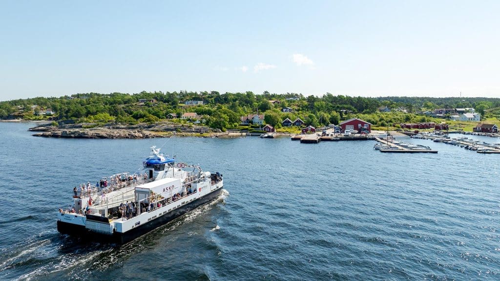 Passasjerferge med folk om bord kjører mot kaia ved Nedgården på Hvaler, omgitt av svaberg og hytter.