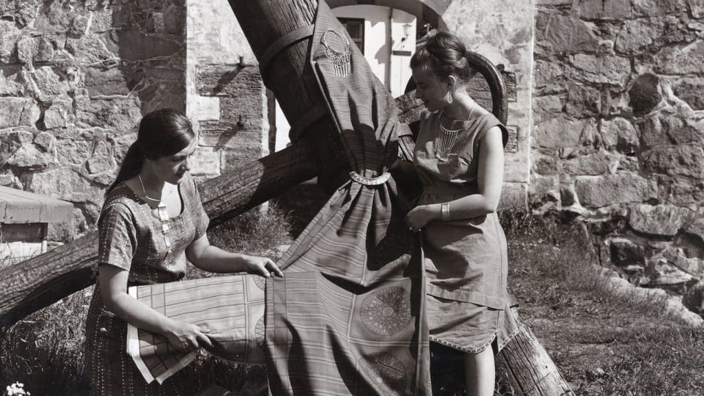 To kvinner viser frem mønstret tekstil hengt over en trestokk foran en gammel steinbygning i Gamlebyen i Fredrikstad. Bildet er fra perioden da kunstnere arbeidet ved PLUS (1958–1978). Til venstre: smykkekunstner Tone Vigeland. Til høyre: tekstilkunstner Kari Fronth.