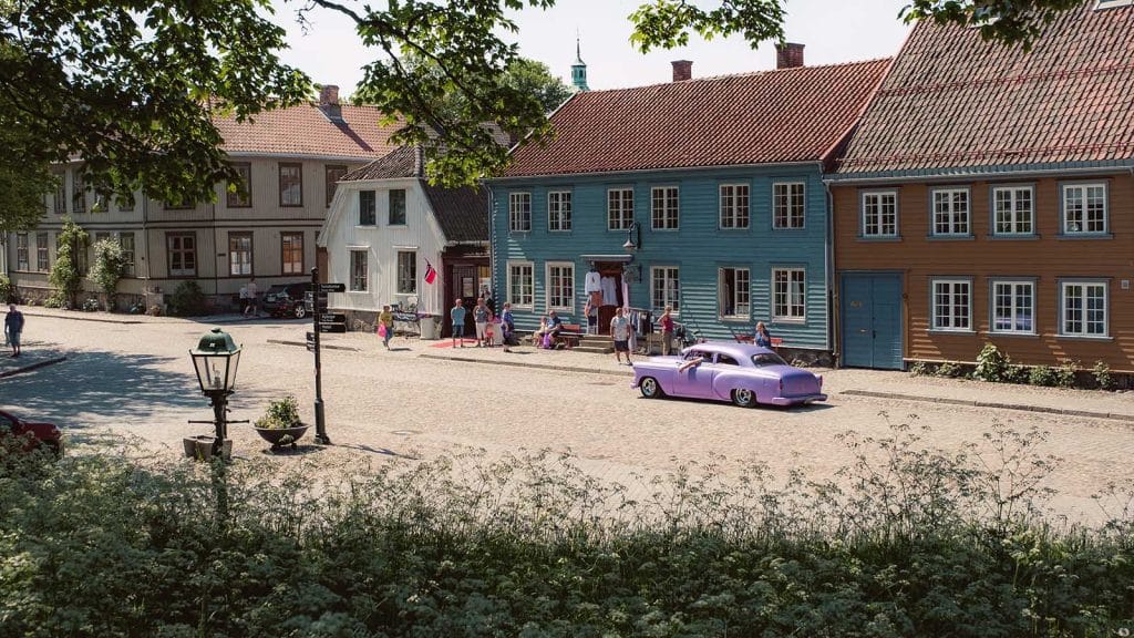 Brosteinsgate i Gamlebyen i Fredrikstad med fargerike trehus, folk som rusler og en lilla veteranbil parkert i solen.