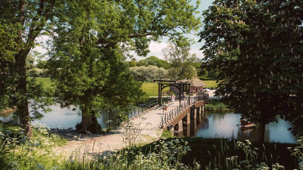 Folk som krysser Vindebrua inn til Gamlebyen i Fredrikstad på en solrik dag, omgitt av grønt landskap og vann.