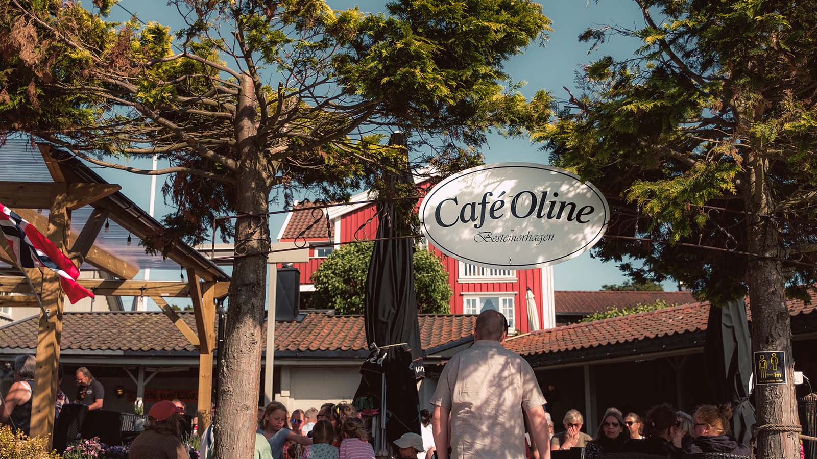 Skilt og uteservering ved Café Oline på Søndre Sandøy, omgitt av trær og folk i sommersol