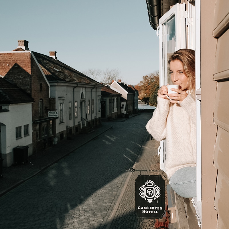 Kvinne sitter i vinduskarmen med kaffekopp og ser utover brosteinsgate i Gamlebyen