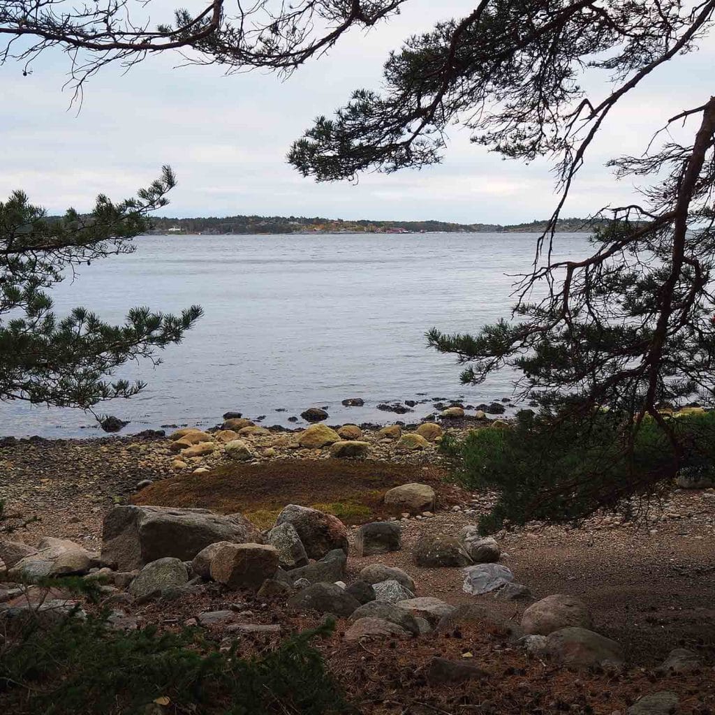Rullesteinstrand og svaberg under furutrær, med utsikt over sjøen.