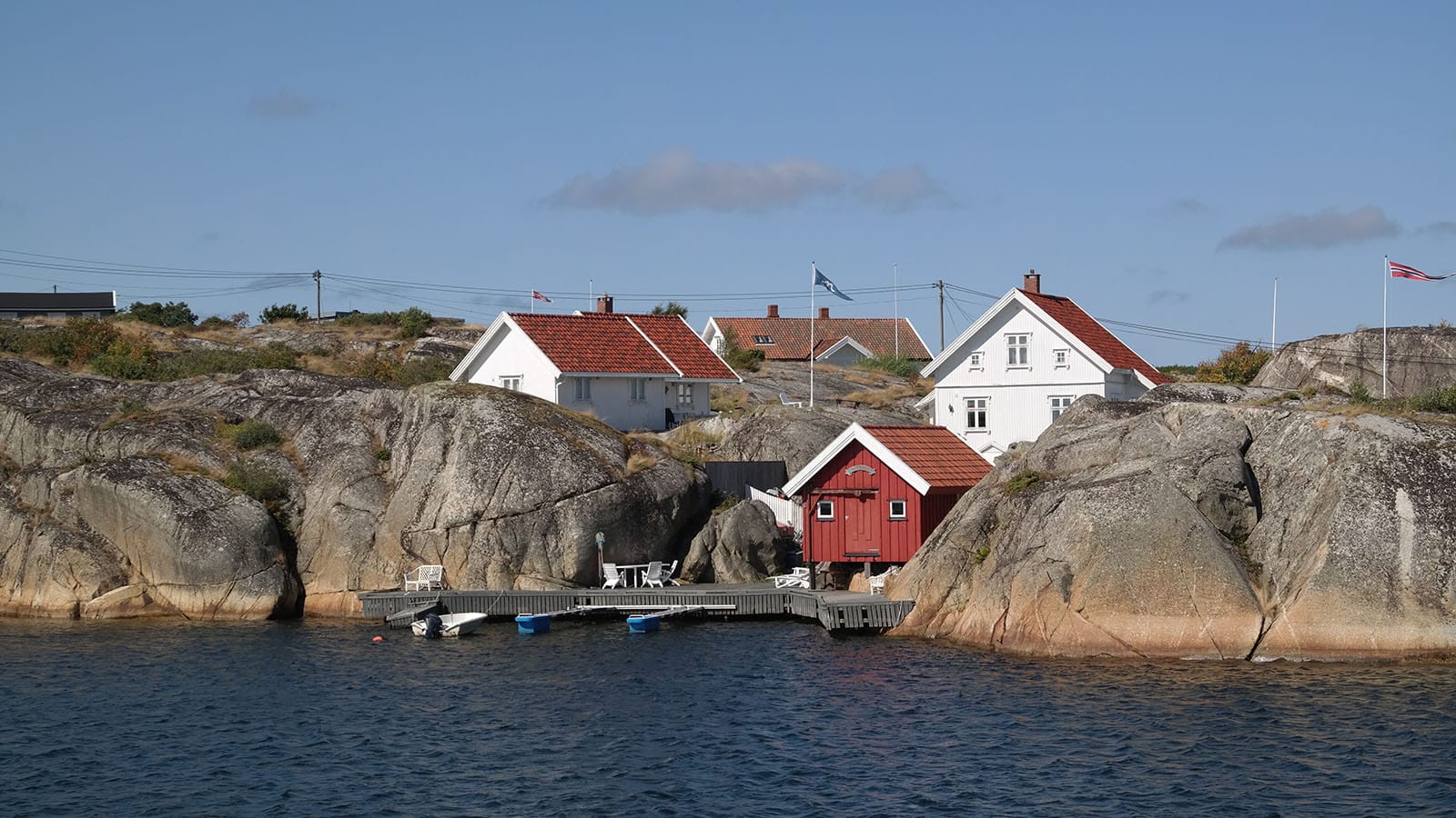 Hvitmalte hus og rød sjøbod mellom svabergene på Lauer, sett fra sjøen, med brygge og småbåter i forgrunnen.