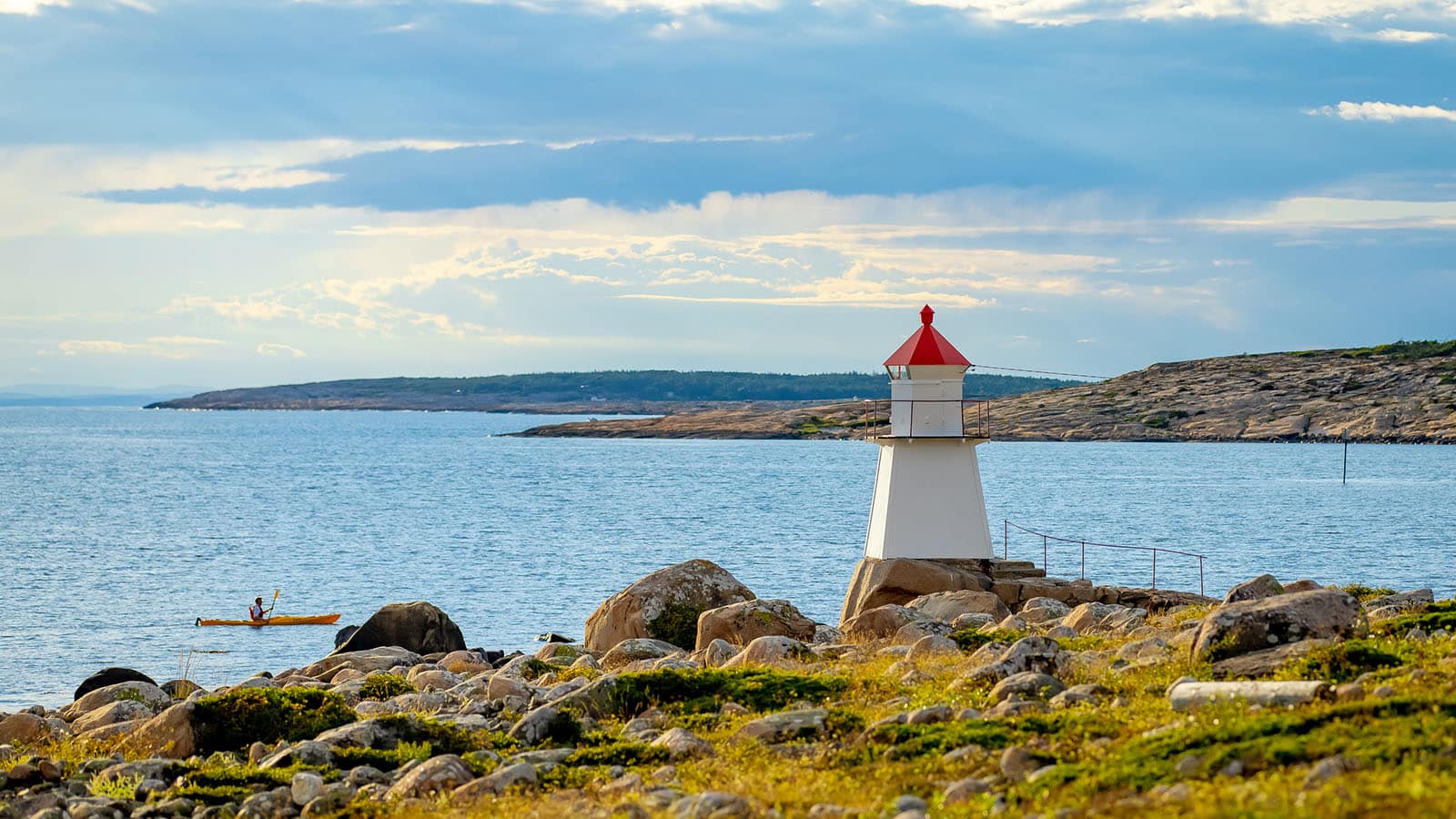 : Kajakkpadler på sjøen ved Pikesten fyrlykt på Asmaløy, omgitt av svaberg og åpent kystlandskap i ettermiddagssol.