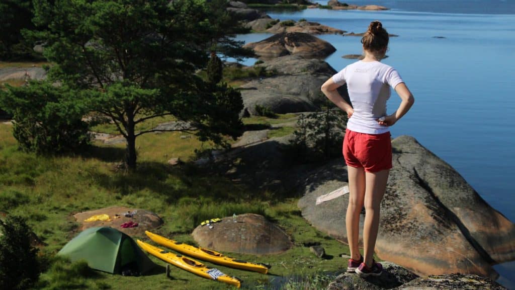 En person skuer utover havet fra en høyde, med telt og kajakker nede på svabergene.