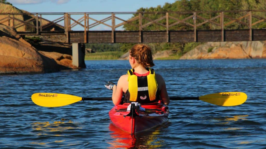 En padler i rød kajakk nærmer seg en trebro i skjærgården, omgitt av klipper og blått vann