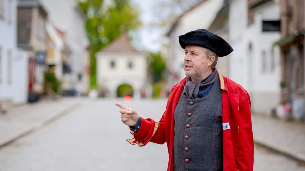 Guide i rød uniform peker og forteller i en brosteinsgate i Gamlebyen i Fredrikstad.