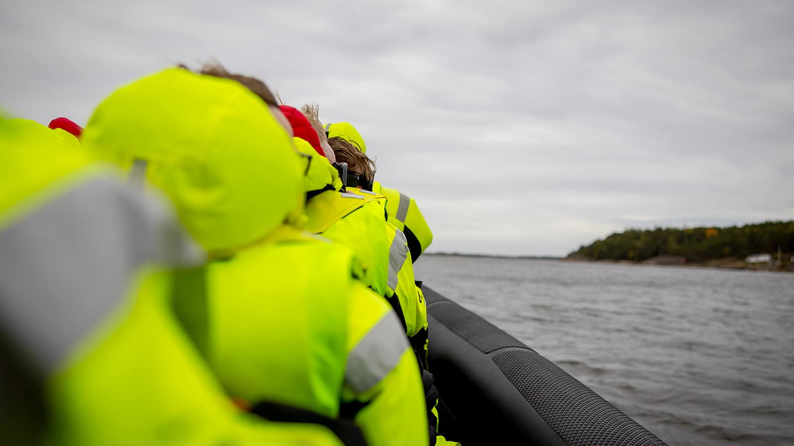 Deltakere i gule drakter sitter tett i RIB-båt, omgitt av grått hav og kystlandskap.